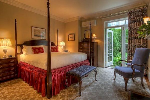 This image shows a cozy bedroom with a four-poster bed, a bench, armchair, lamps, and a dresser, with open doors leading to a garden area.