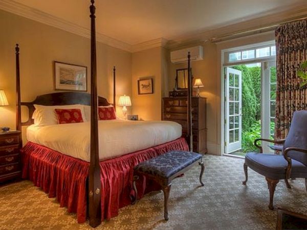 A cozy Newport, RI, guest room with a king-size bed, a bench, armchair, lamps, a dresser, and doors leading to a garden patio.