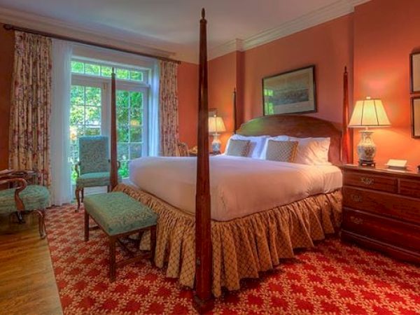 A cozy Newport, RI, guest room with a king-size bed, a bench, 2 armchairs, lamps, a dresser, and doors leading to a garden patio.