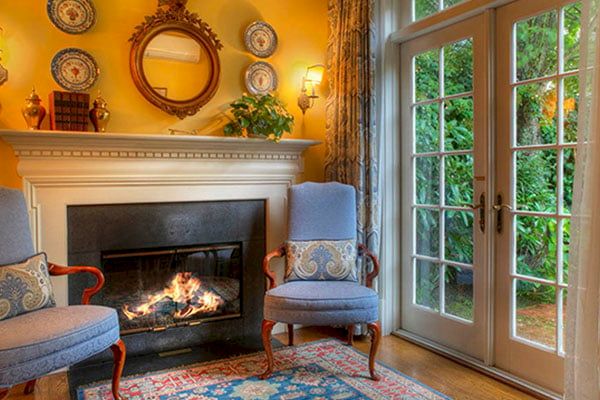 A cozy room features a lit fireplace, two armchairs, a French door with garden view, and decorative plates on mantels above, lending a warm ambiance.