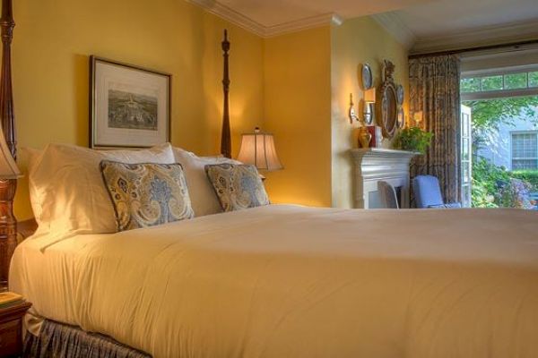 A cozy bedroom with a large bed, decorative pillows, yellow walls, wall art, lamps, a white mantle, mirrors, and a view through a window to the outside.