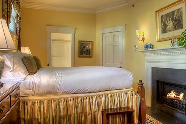 A cozy bedroom with a neatly made bed, skirted bedding, a fireplace, framed artwork on the walls, and a door leading to another room ending the sentence.