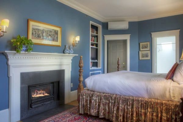 A cozy bedroom with blue walls features a fireplace, a large bed with a skirted base, bookshelf, wall art, and an air conditioning unit.