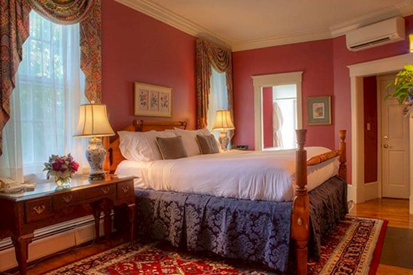 The image shows a cozy bedroom with a four-poster bed, bedside lamps, a wooden desk with a chair, a rug, and decor including paintings and curtains.