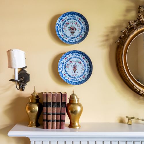 A decorative mantel with two blue plates on the wall, a round mirror, a candle sconce, brass vases, and a set of books.