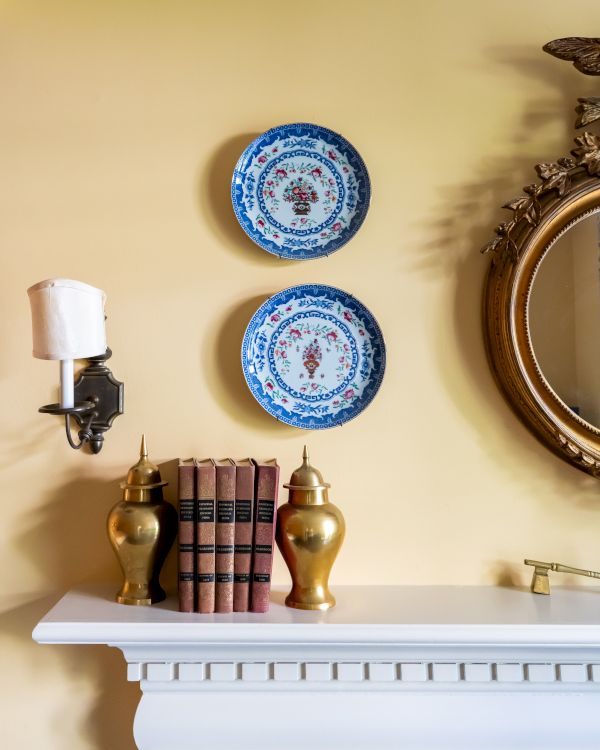 A decorative mantel with two blue plates on the wall, a round mirror, and a set of books at The Francis Malbone House.