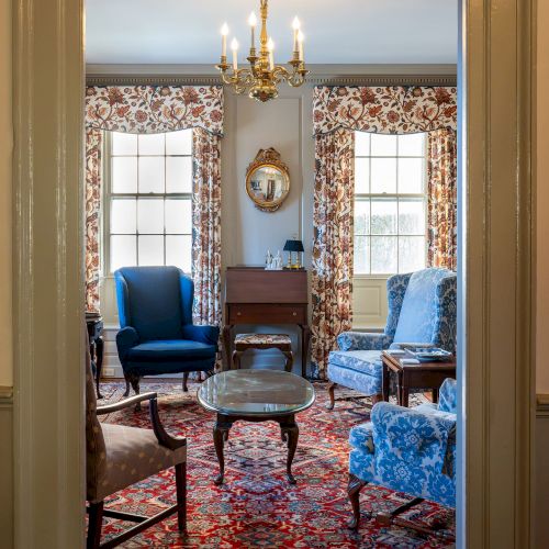 A common space at The Francis Malbone House featuring three blue arm chairs, a fireplace, and bright windows with floral curtains.