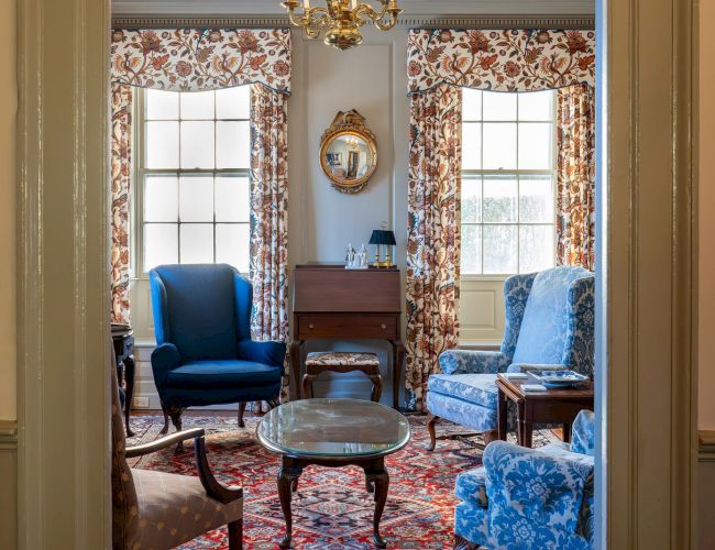 A cozy living room with upholstered chairs, a coffee table, a chandelier, floral curtains, a rug, and a small desk, viewed through a doorway.