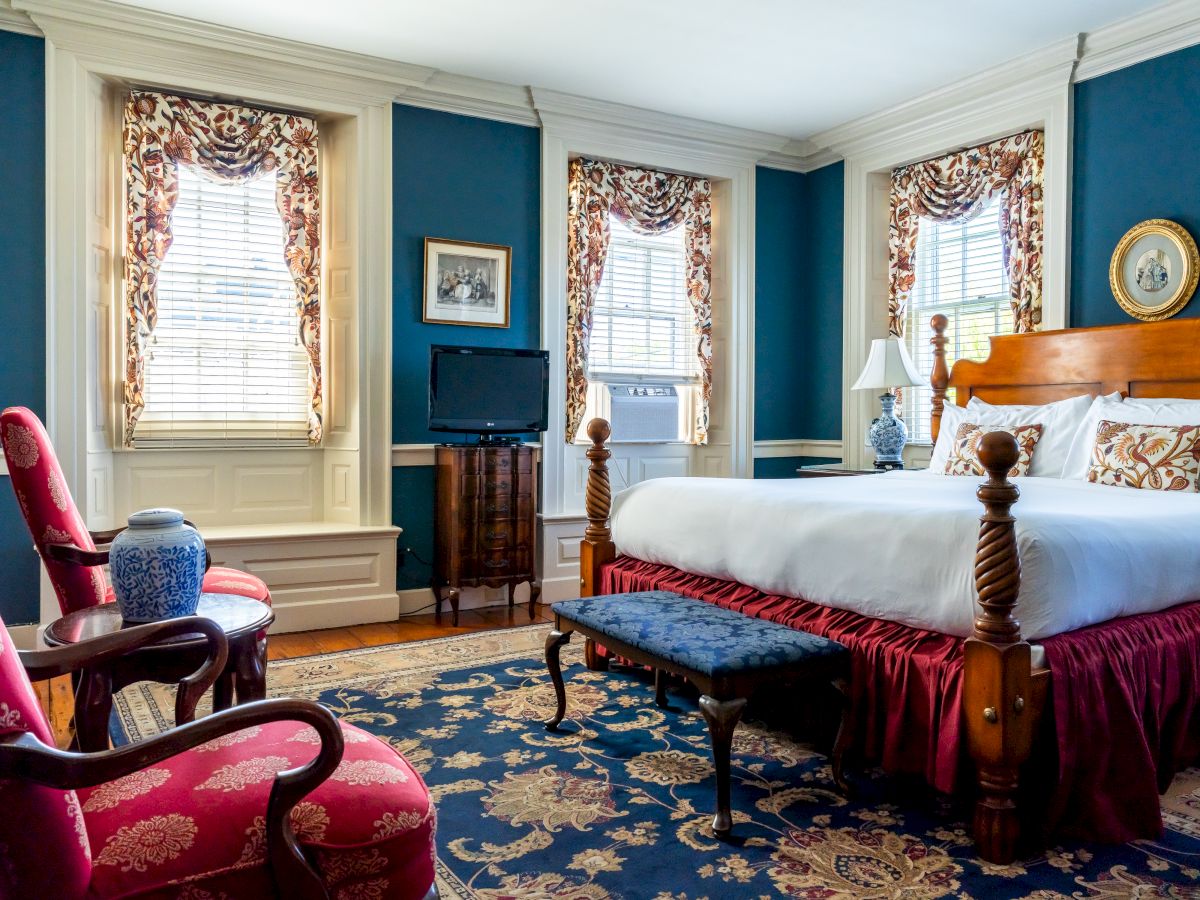 A well-furnished bedroom with blue walls, a large wooden bed, two red armchairs, a dresser with a TV, and three windows with patterned curtains.