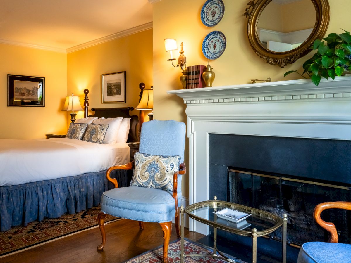 The image shows a cozy bedroom with a large bed, blue chairs, a fireplace, framed artwork, a mirror, decorative plates, and a plant.