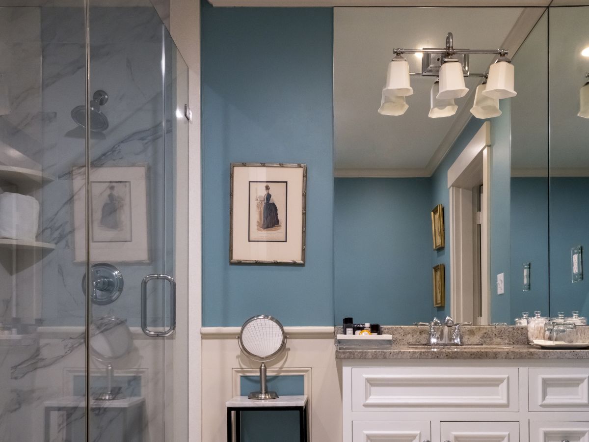 A bathroom with blue walls, a glass shower, a sink with a mirror, and wall-mounted light fixtures. There are framed artworks and a vanity table.