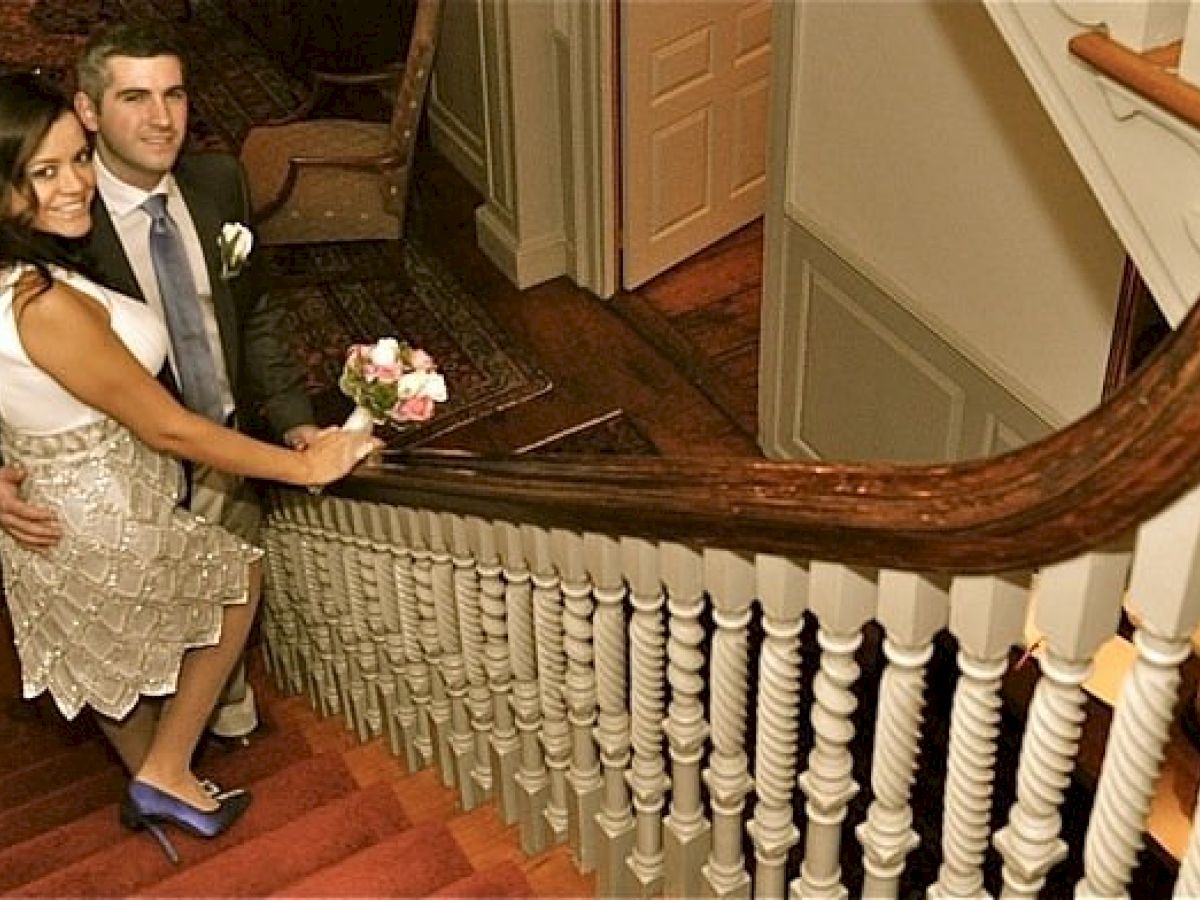 A couple in formal attire stands on a staircase, with the woman holding a bouquet of flowers, looking up at the camera.