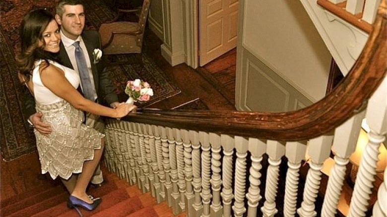 A couple in formal attire stands on a staircase, with the woman holding a bouquet of flowers, looking up at the camera.