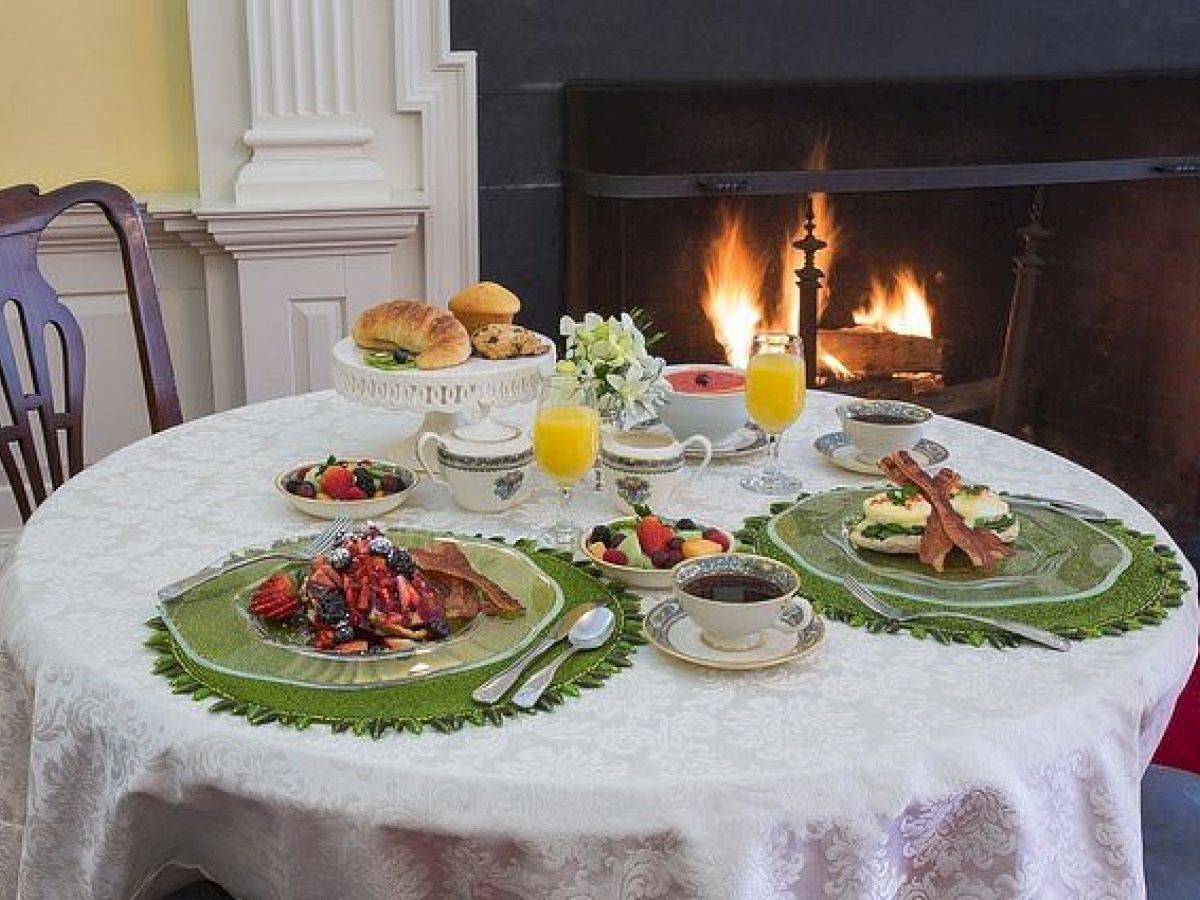 A table set for breakfast with plates of fruits, waffles, bacon, juice, and coffee near a fireplace at The Francis Malbone House.