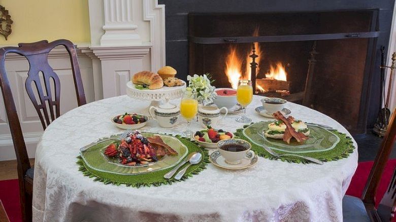 A table set for breakfast with plates of fruits, waffles, and bacon, alongside orange juice and coffee, near a cozy fireplace.