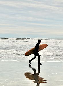 A person in a wetsuit is walking along the beach carrying a surfboard, with the ocean and waves in the background.