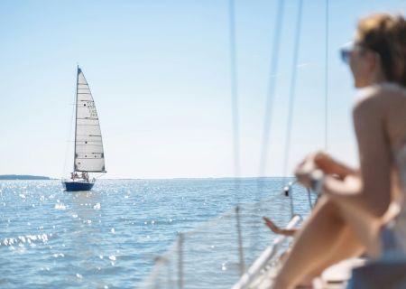 A person on a boat gazes at a sailing yacht on a sunny, calm sea, creating a serene and picturesque scene.