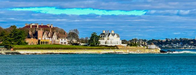 Large, elegant houses on a coastal landscape with a bright blue ocean in the foreground and a partly cloudy sky in the background.