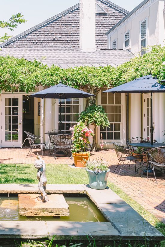 A charming courtyard featuring a small fountain, surrounded by tables with blue umbrellas and lush greenery, providing a serene outdoor setting.