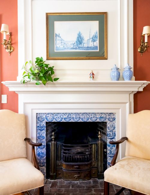 A gas fireplace with blue and white tiles, flanked by two armchairs with a framed painting, a plant, and two vases on the mantel.