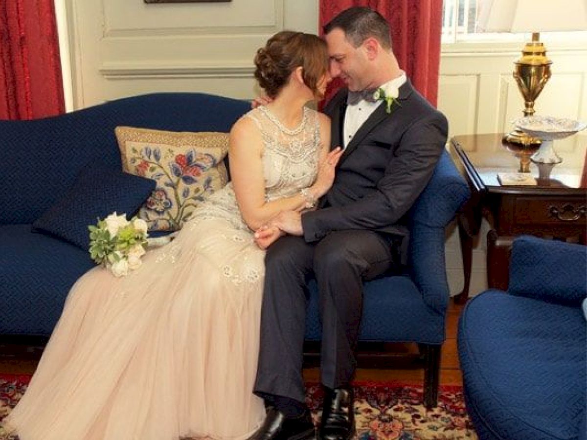A couple dressed in wedding attire sit closely together on a sofa with floral pillows in an elegantly decorated room.