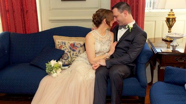 A couple dressed in wedding attire is sitting close together on a blue sofa, sharing an intimate moment and holding a bouquet of flowers.