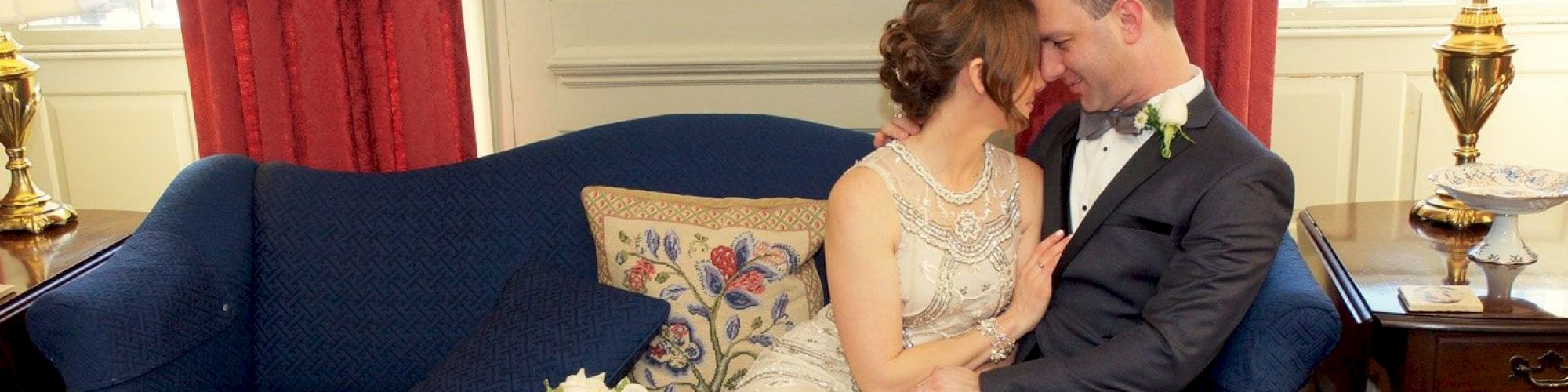 A couple, dressed in wedding attire, sits closely embracing on a blue couch with floral pillows, in a room with red curtains and elegant decor.