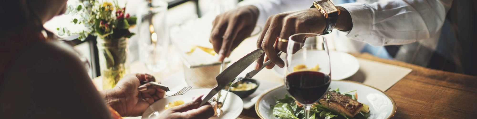 Two people are dining at a restaurant, enjoying a meal with wine. One is cutting food on a plate, and the table is set elegantly.