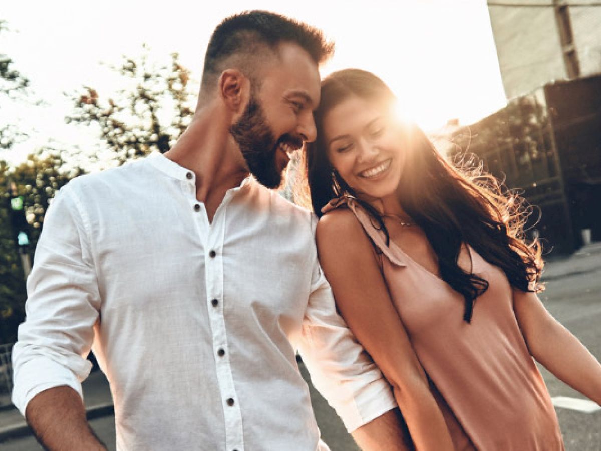 A man in a button-up shirt and a woman in a pink blouse walk together outdoors holding hands with the sun setting behind them.