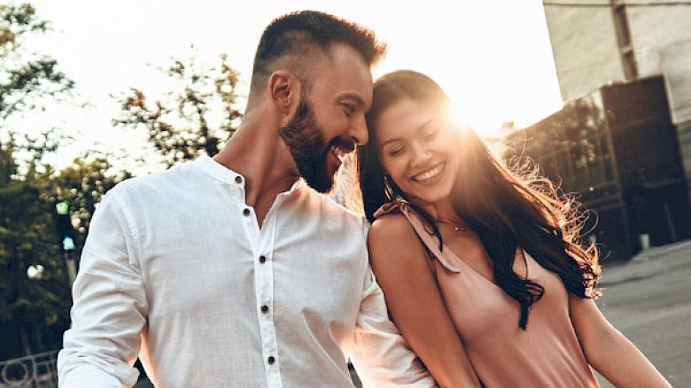 A smiling couple walks outdoors, hand in hand, with the sun setting behind them. They appear happy and engaged in each other's company.