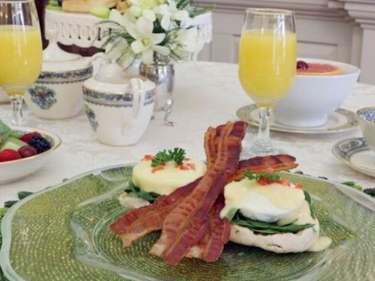 A breakfast table with Eggs Benedict topped with bacon, fruit salad, floral centerpiece, and glasses of orange juice.