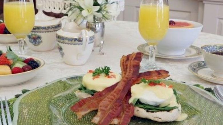 A breakfast table with Eggs Benedict topped with bacon, fruit salad, floral centerpiece, and glasses of orange juice ending the sentence.