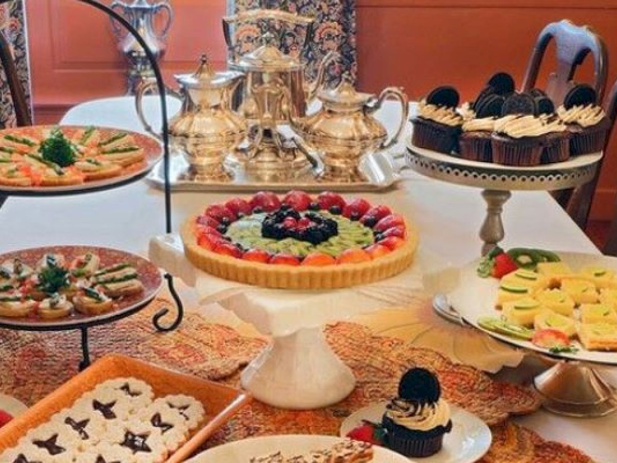 A table setup featuring assorted pastries, tarts, cupcakes, and silver tea sets ready for a high tea or dessert gathering.