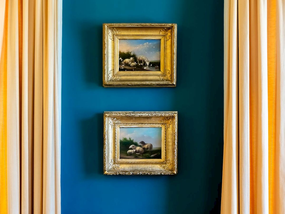 The image shows two framed paintings on a blue wall, flanked by beige curtains, with a white wall paneling below.