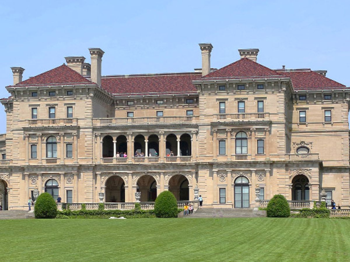 A large, historic mansion with a green lawn in front, characterized by multiple arches, windows, and chimneys, under a clear sky.