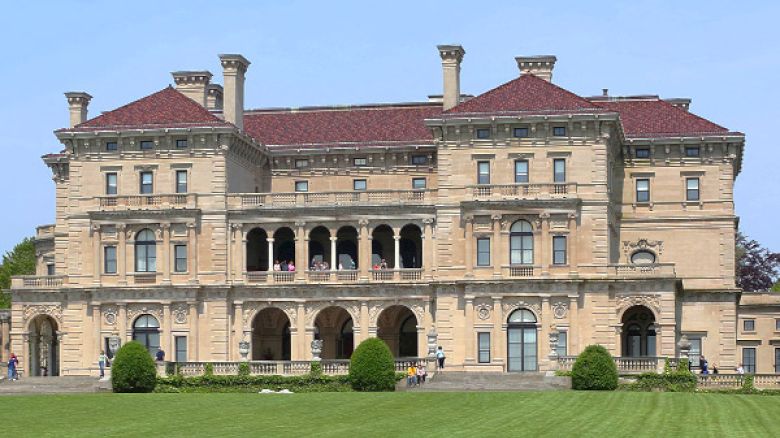 This image shows a large, historic mansion with a green lawn in front, characterized by multiple arches, windows, and chimneys, under a clear sky.