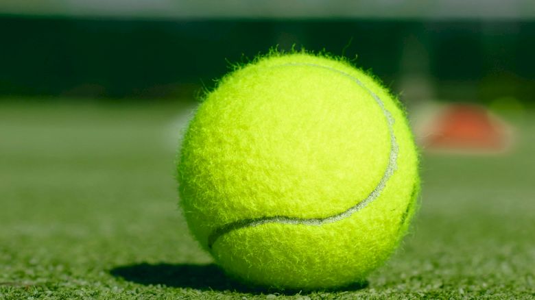 A tennis ball resting on a grass court with a blurry background, likely suggesting an outdoor tennis court.