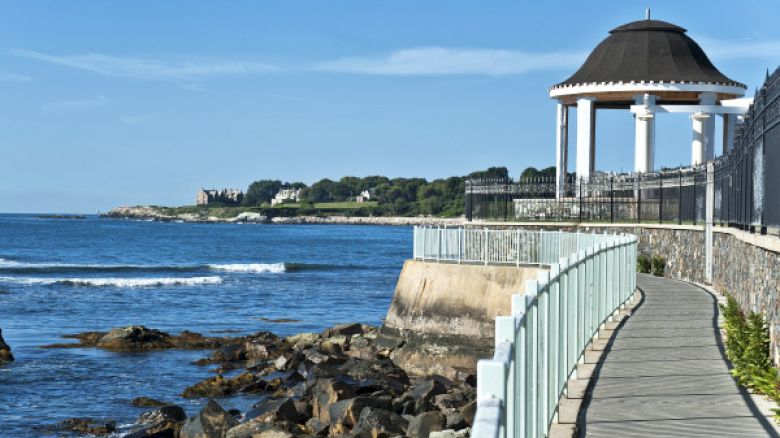 A seaside walkway with a coastal gazebo, stone wall, and a distant mansion on a sunny day by the ocean.