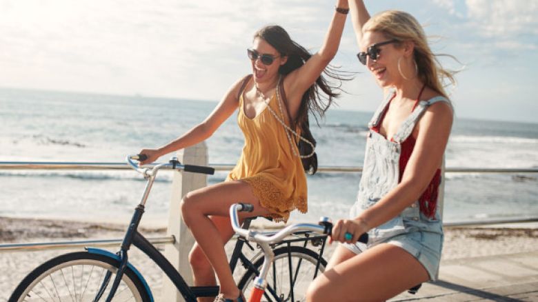 Two women riding bicycles by the ocean, holding hands and smiling, under a clear sky with the sea in the background.