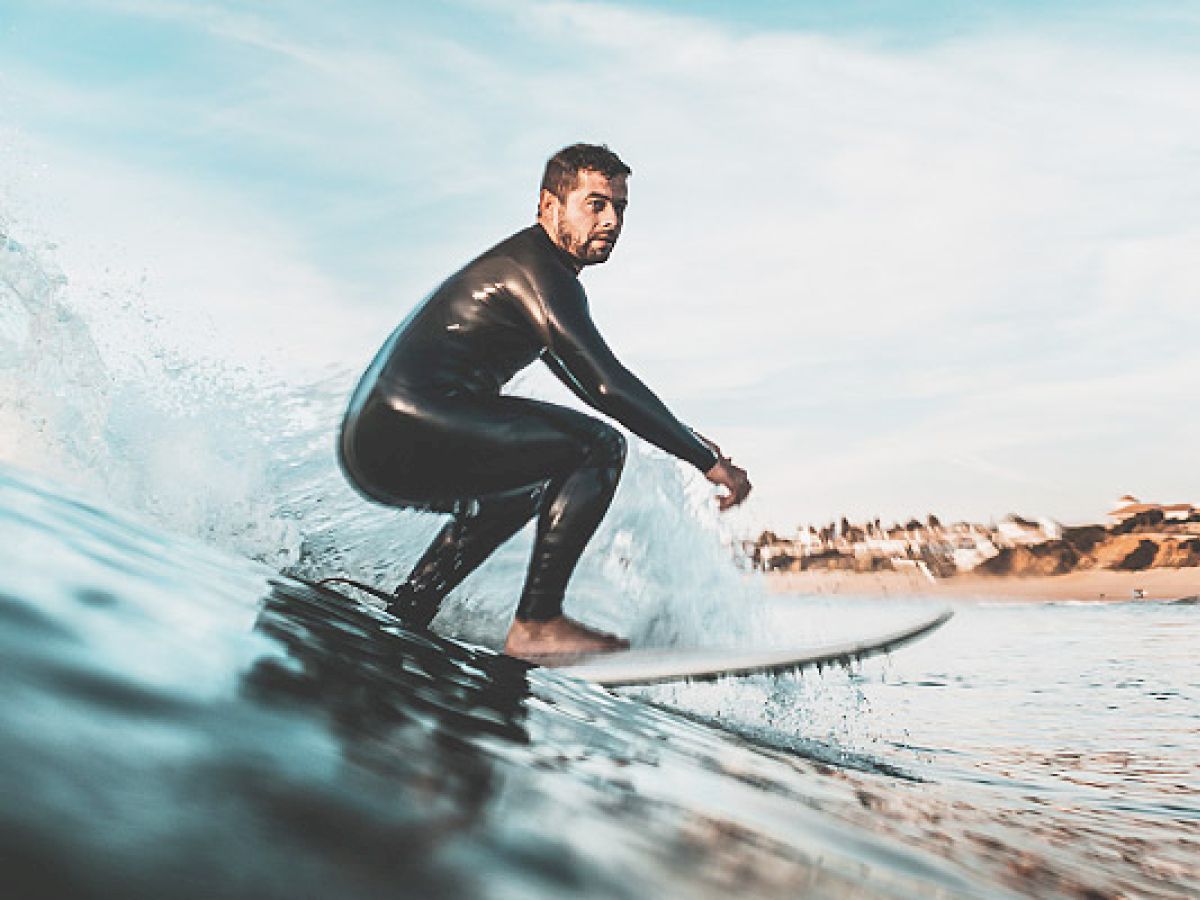 A person in a wetsuit is surfing on a wave with a coastline and buildings visible in the background, enjoying the water.