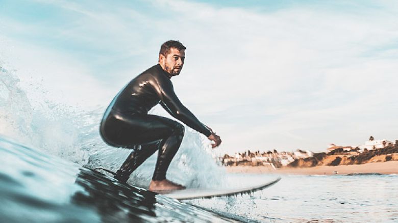 A person in a wetsuit is surfing on a wave with a coastline and buildings visible in the background, enjoying the water.