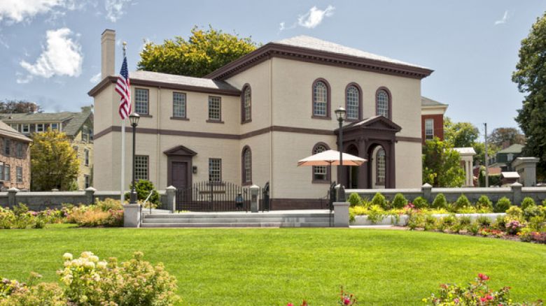 A historic two-story building with arched windows, surrounded by a well-maintained garden, an American flag, and an umbrella-covered seating area.