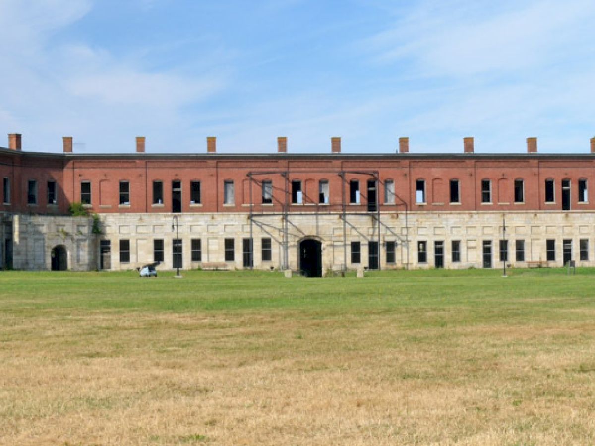 The image shows a historic brick and stone building with multiple windows, a central archway, and an open grassy area in front.