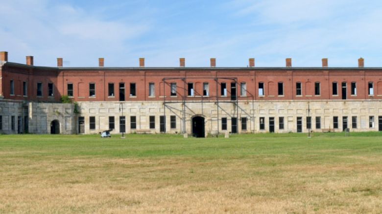 The image shows a historic brick and stone building with multiple windows, a central archway, and an open grassy area in front.