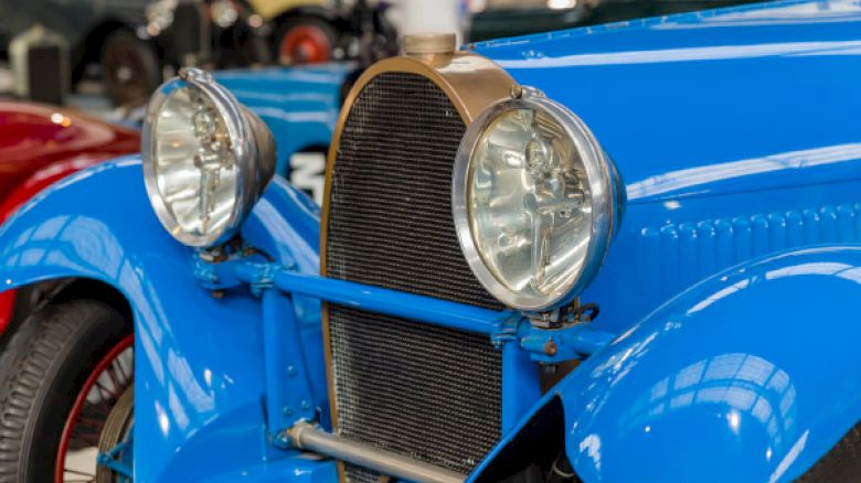 A close-up of the front end of a vintage blue car, showcasing its headlights, radiator grille, and classic lines, with other vehicles in the background.