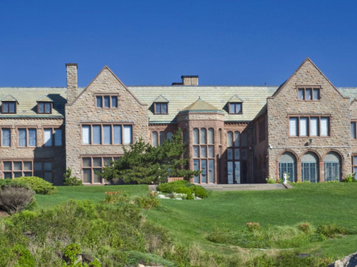 The image depicts a large, grand stone mansion with multiple windows and green lawns, set against a clear blue sky.