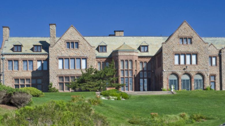 The image depicts a large, grand stone mansion with multiple windows and green lawns, set against a clear blue sky.