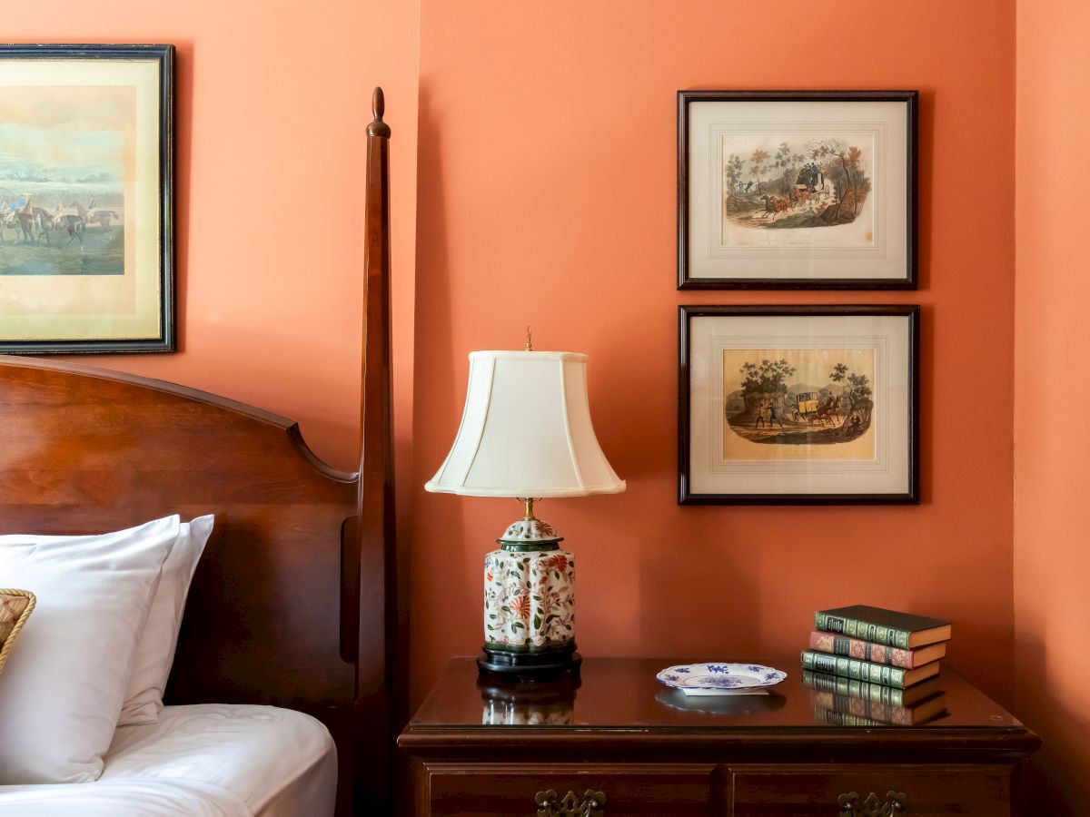 A cozy bedroom corner with a wooden bed, vintage lamp, stacked books, a decorative plate, and framed artwork on a peach-colored wall.