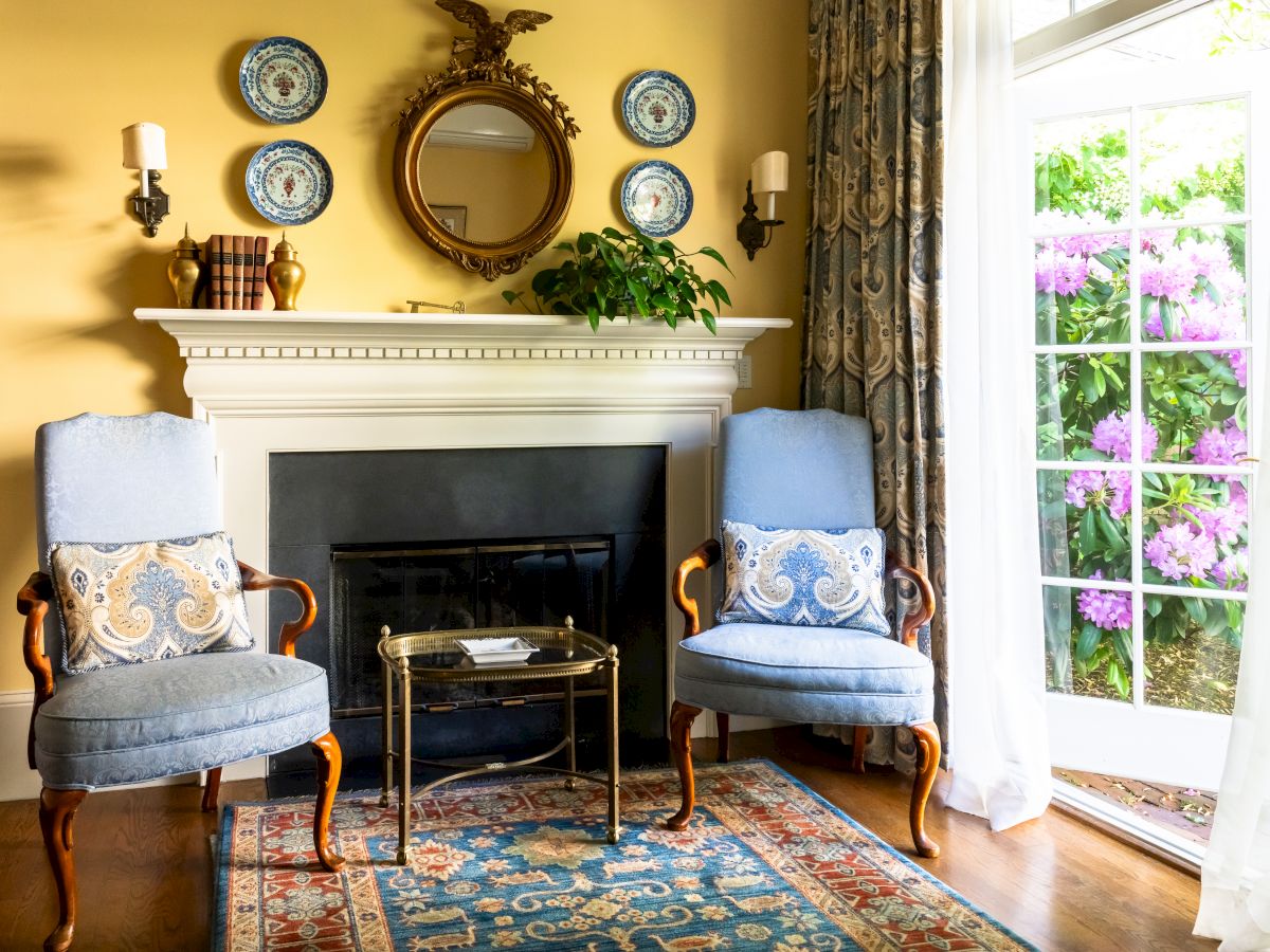 A cozy living room with blue chairs, a decorated fireplace, a small table, and a rug, situated near large windows letting in natural light.