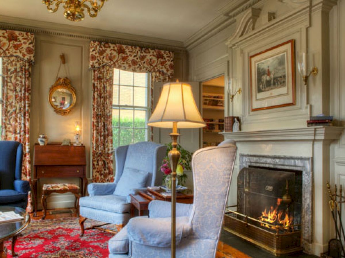 A common space at The Francis Malbone House featuring three blue arm chairs, a fireplace, and bright windows with floral curtains.